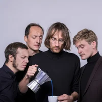 Ein Bandmitglied von Morland Shaker gießt sich aus einer Espressokanne eine blaue Flüssigkeit in eine Espressotasse. Die drei anderen Musiker – alle tragen schwarze Rollkragenpullis - schauen kritisch auf das Geschehen.